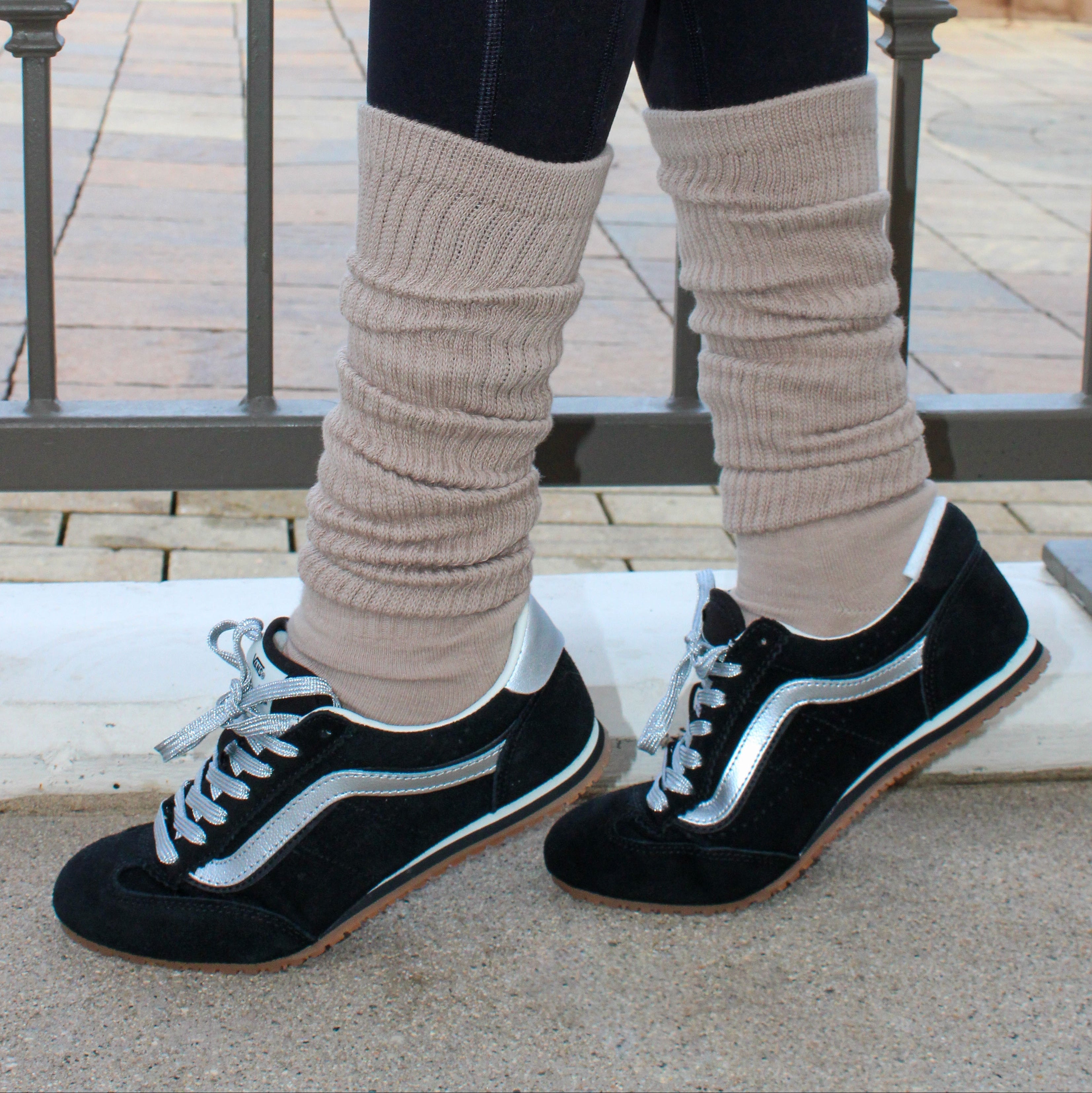 Person wearing black sneakers with white soles, latte colored tici active  slouch socks, and dark pants on a stone surface.