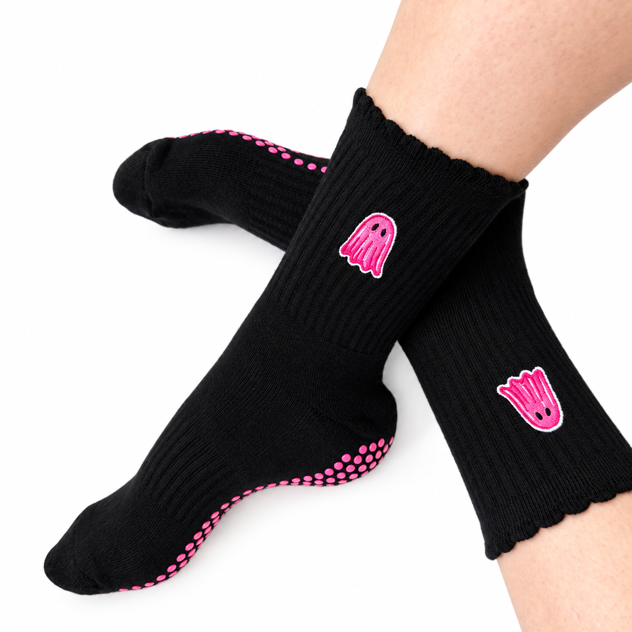 A pair of black crew socks worn on feet, featuring pink ghost graphics and pink grip dots on the sole, against a plain white background.