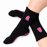 A pair of black crew socks worn on feet, featuring pink ghost graphics and pink grip dots on the sole, against a plain white background.