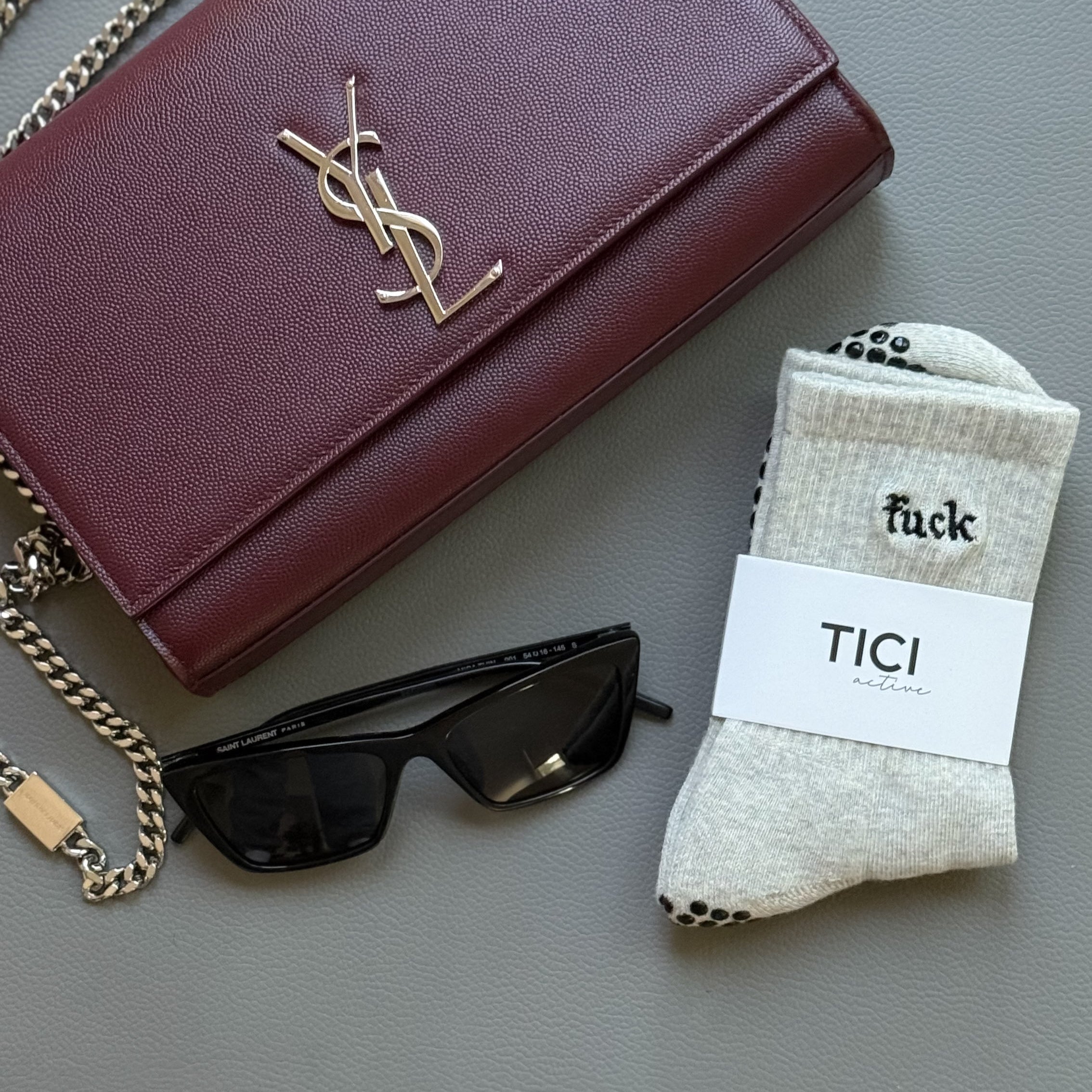 Maroon YSL handbag with chain, YSL sunglasses, and a pair of TICI ACTIVE grip socks on a light gray surface. Socks reads FUCK in black embroidery thread.