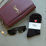 Maroon YSL handbag, YSL sunglasses, and black socks TICI ACTIVE grip socks with black grips with a red heart logo on a light surface.