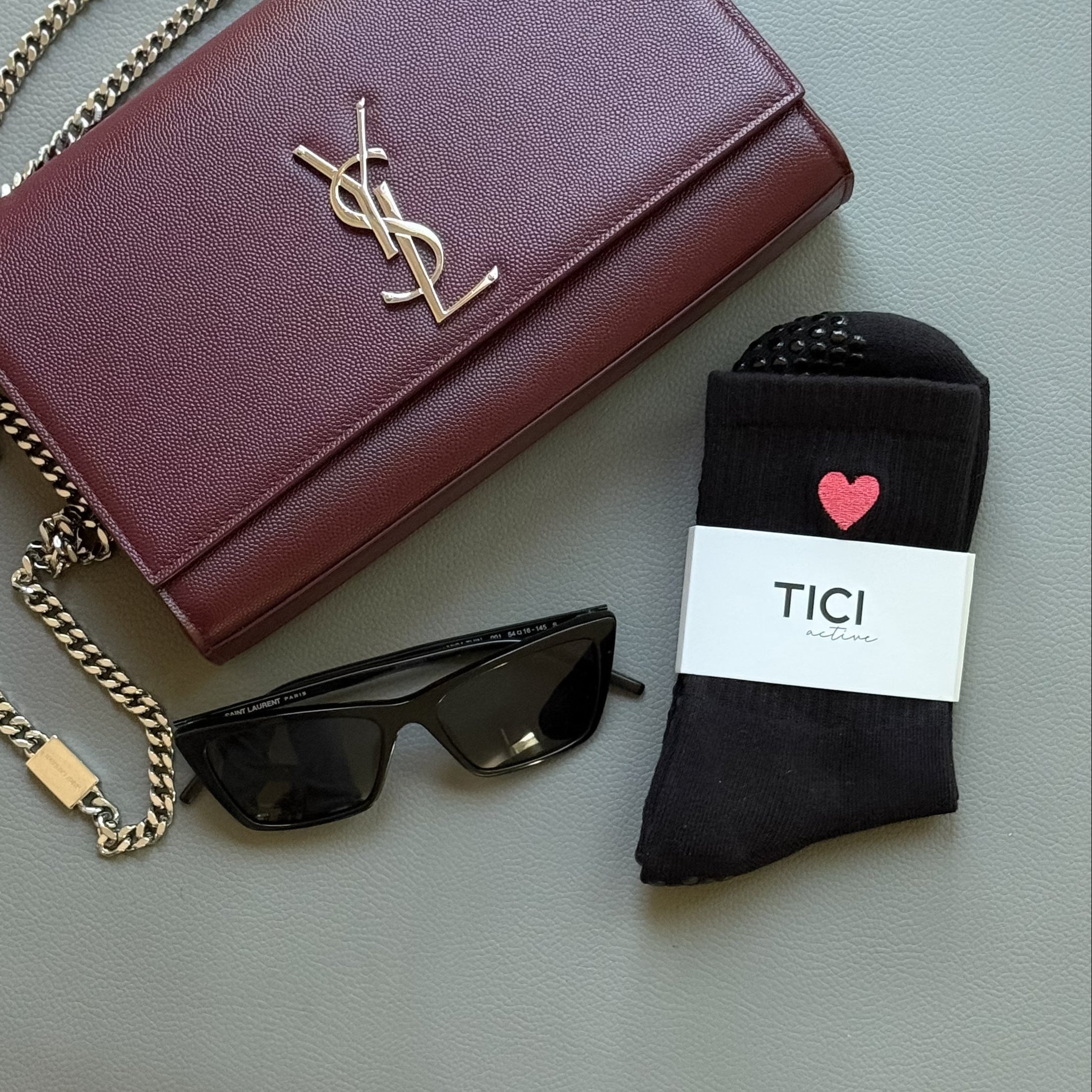Maroon YSL handbag, YSL sunglasses, and black socks TICI ACTIVE grip socks with black grips with a red heart logo on a light surface.