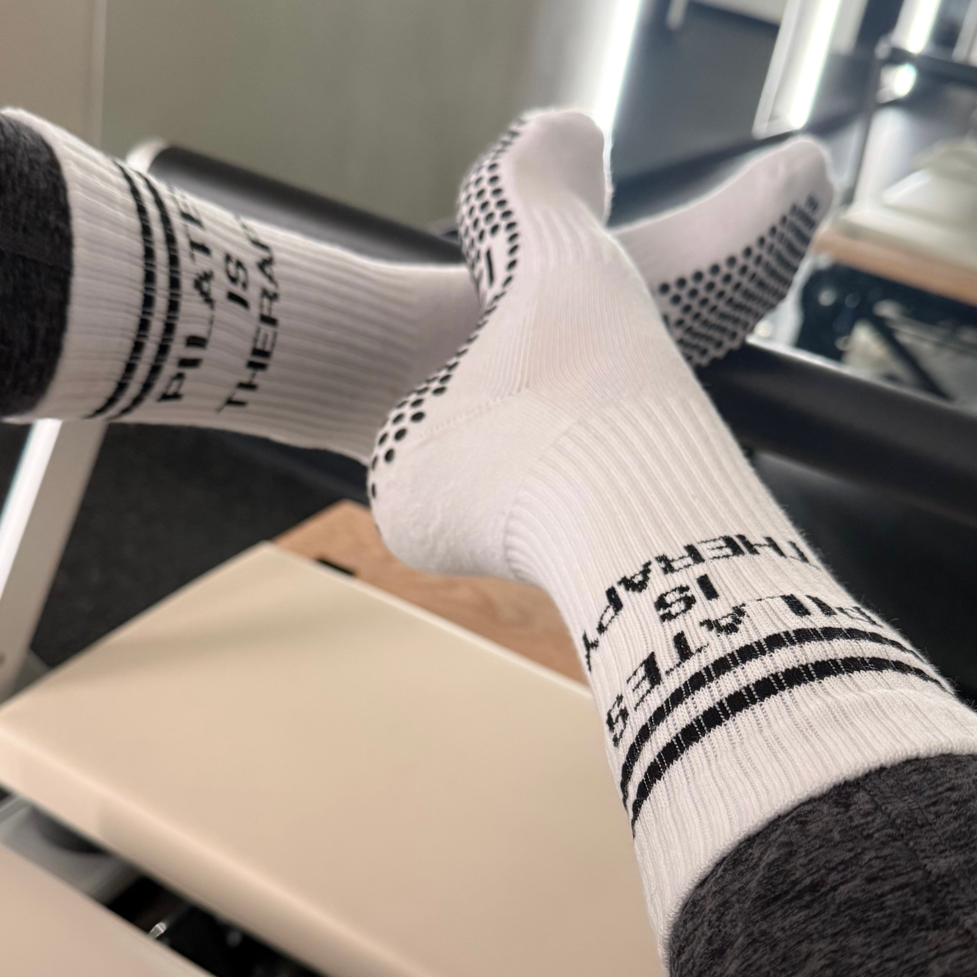 Person wearing white pilates socks with black grips with text that reads, "pilates is therapy", sitting on a pilates reformer.
