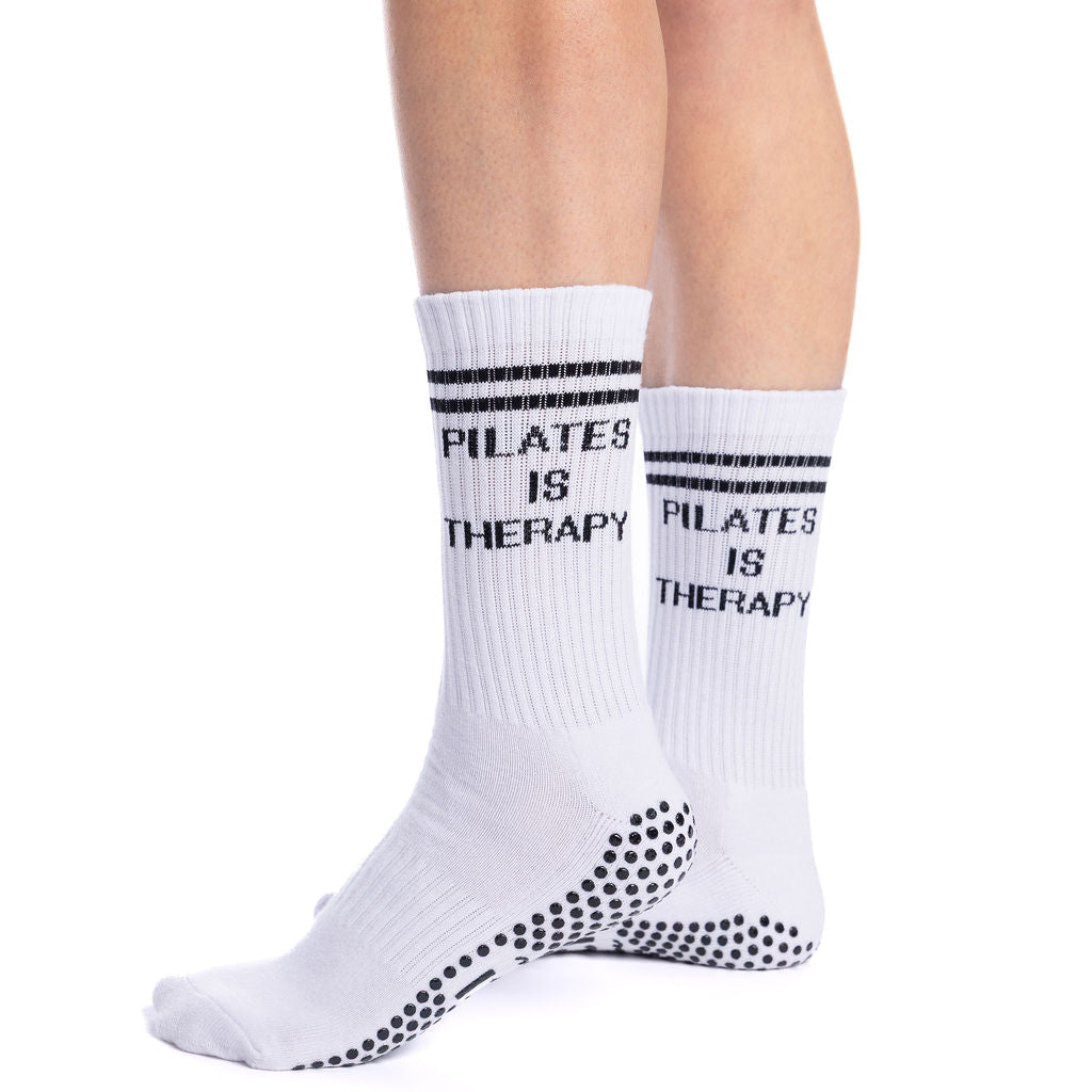 White socks with 'Pilates is Therapy' text worn by a person on a white background